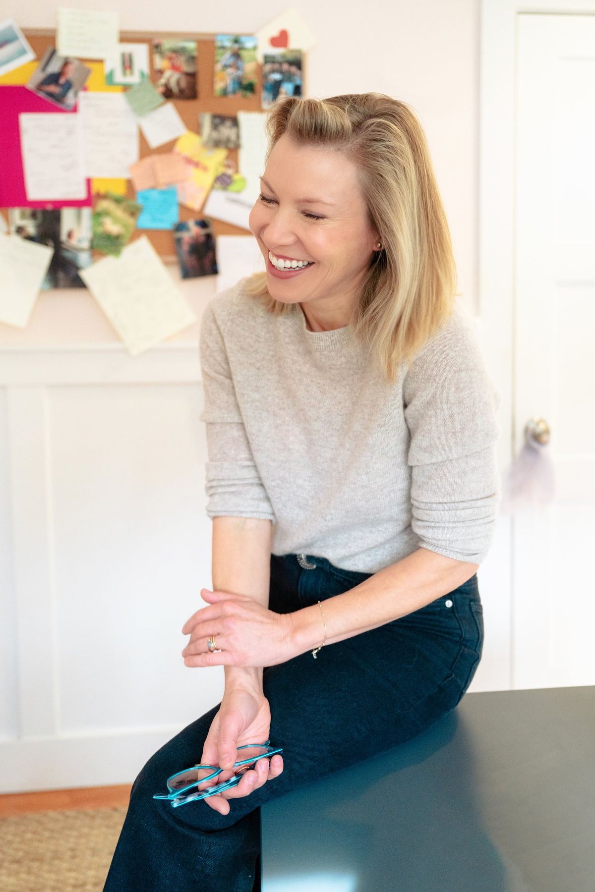 A woman in a soft grey sweater laughs as she perches on a desk in front of a pinboard of notes and photos, glasses in hand