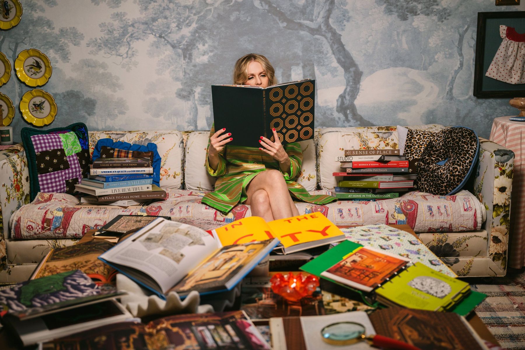 Woman on a sofa peeking over a book, a coffee table piled high with books and magazines in front of her