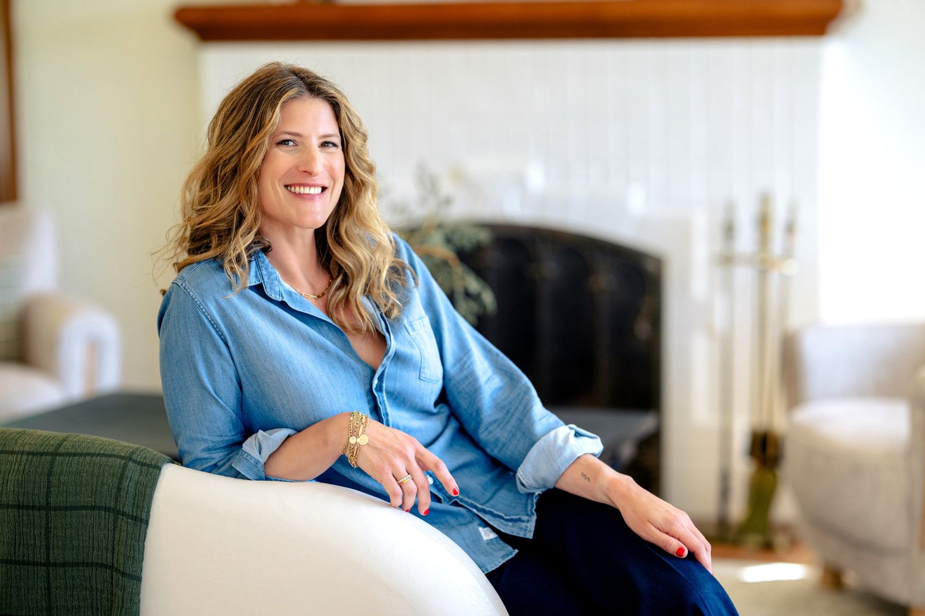 Smiling woman in a chambray shirt relaxing in a white armchair by a fireplace