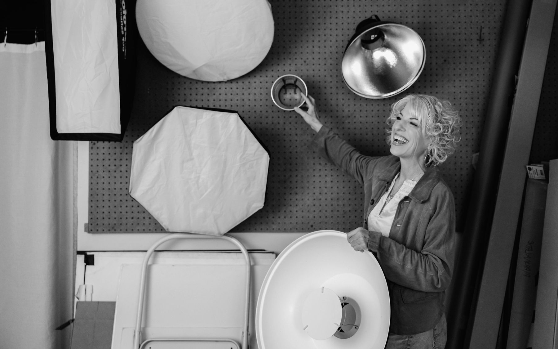Black-and-white photo of Suzanne laughing as she adjusts lighting gear on her studio pegboard wall