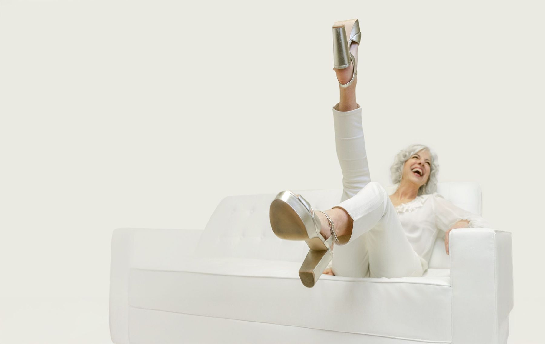 Suzanne laughing on a white sofa with one gold platform heel kicked high in the air