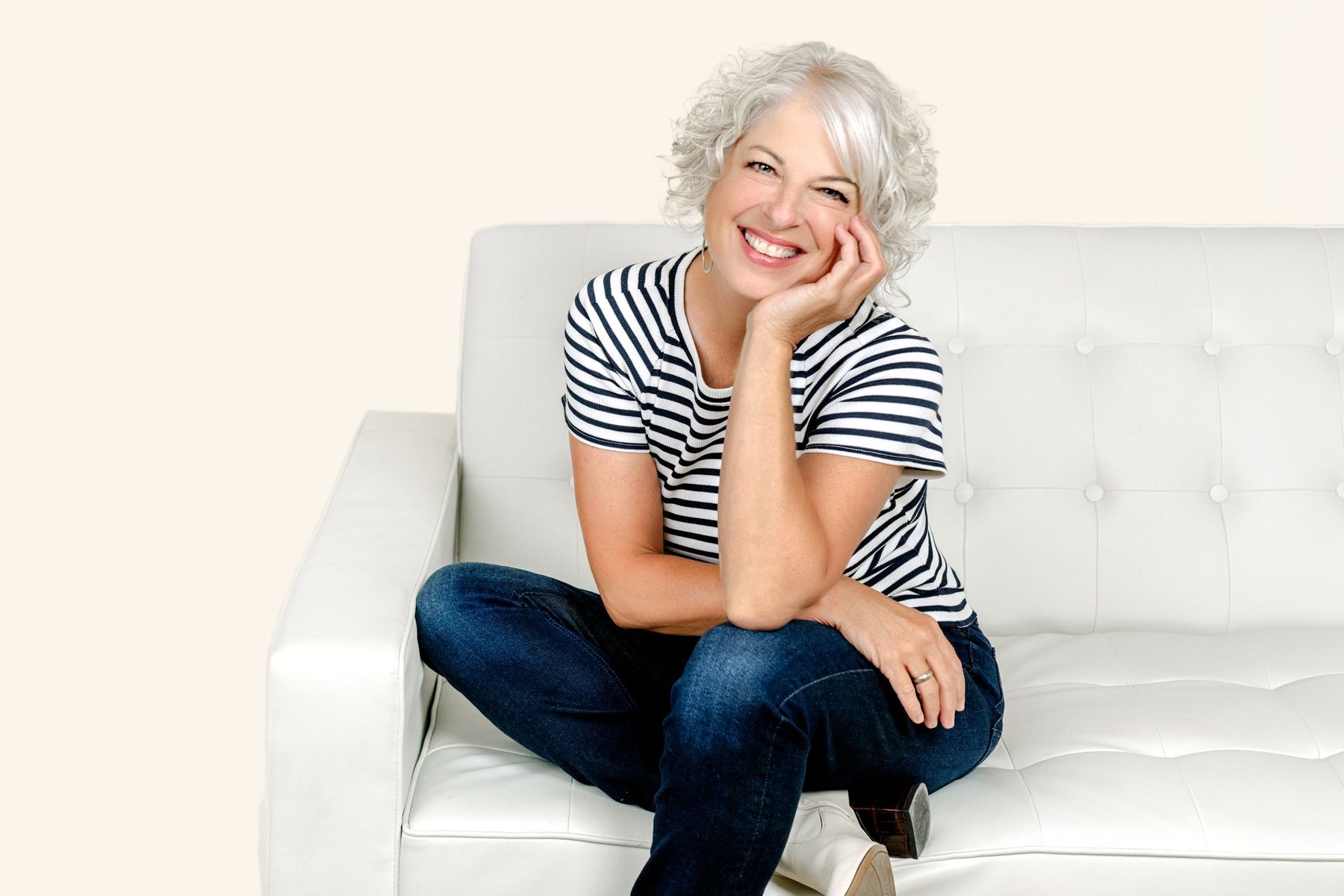 Suzanne smiling, chin on hand, seated on a white tufted sofa in a striped tee and jeans