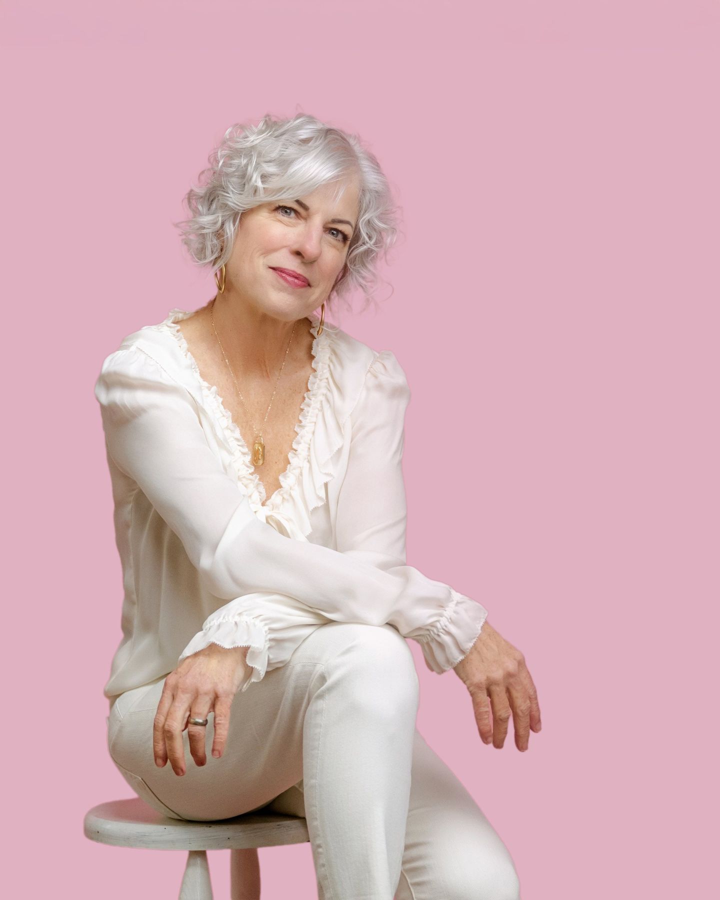 Suzanne Rothmeyer, silver-haired and smiling, seated on a stool in white against a pink backdrop