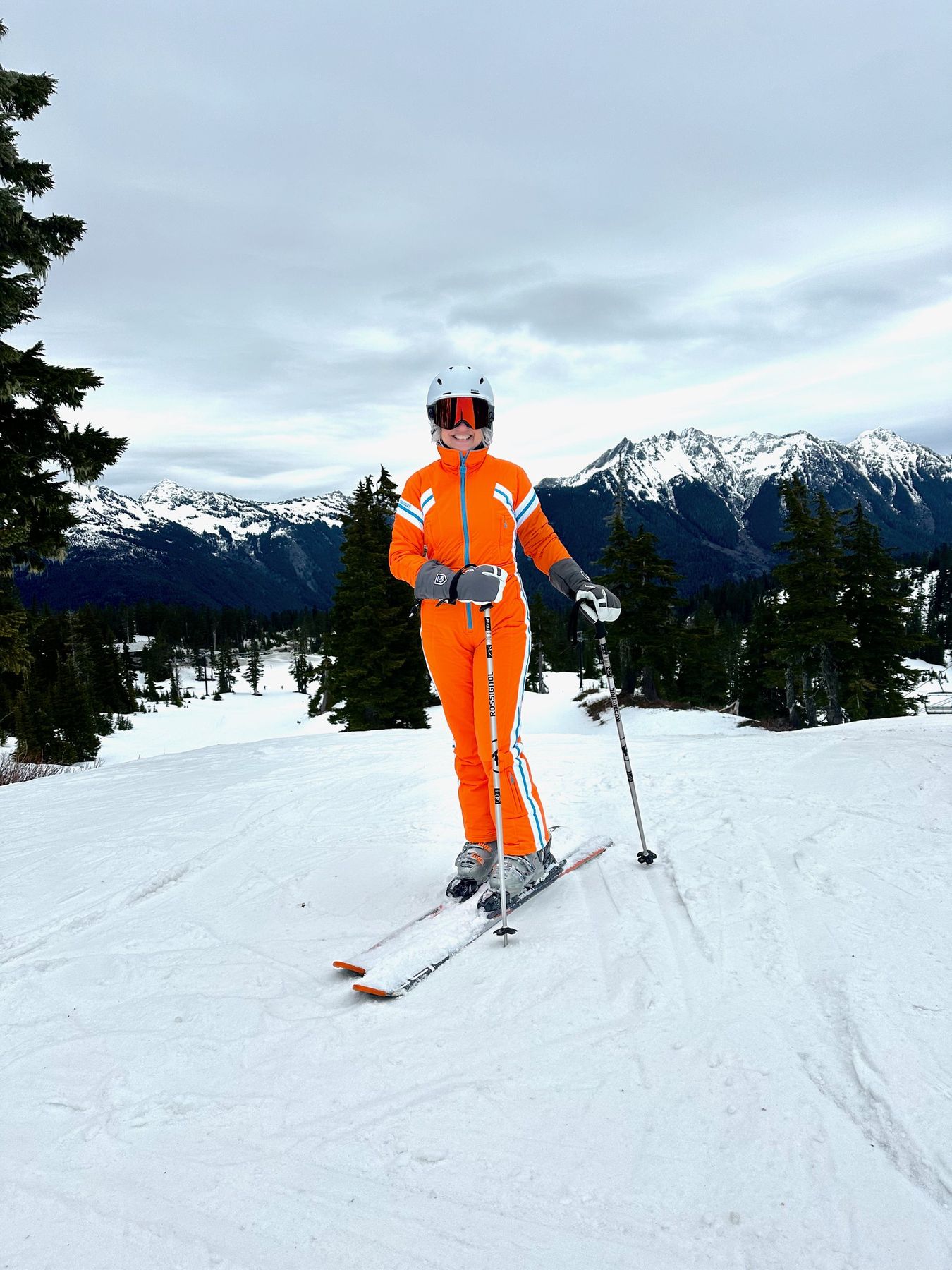 Suzanne skiing in a bright orange snowsuit with snowy peaks behind her