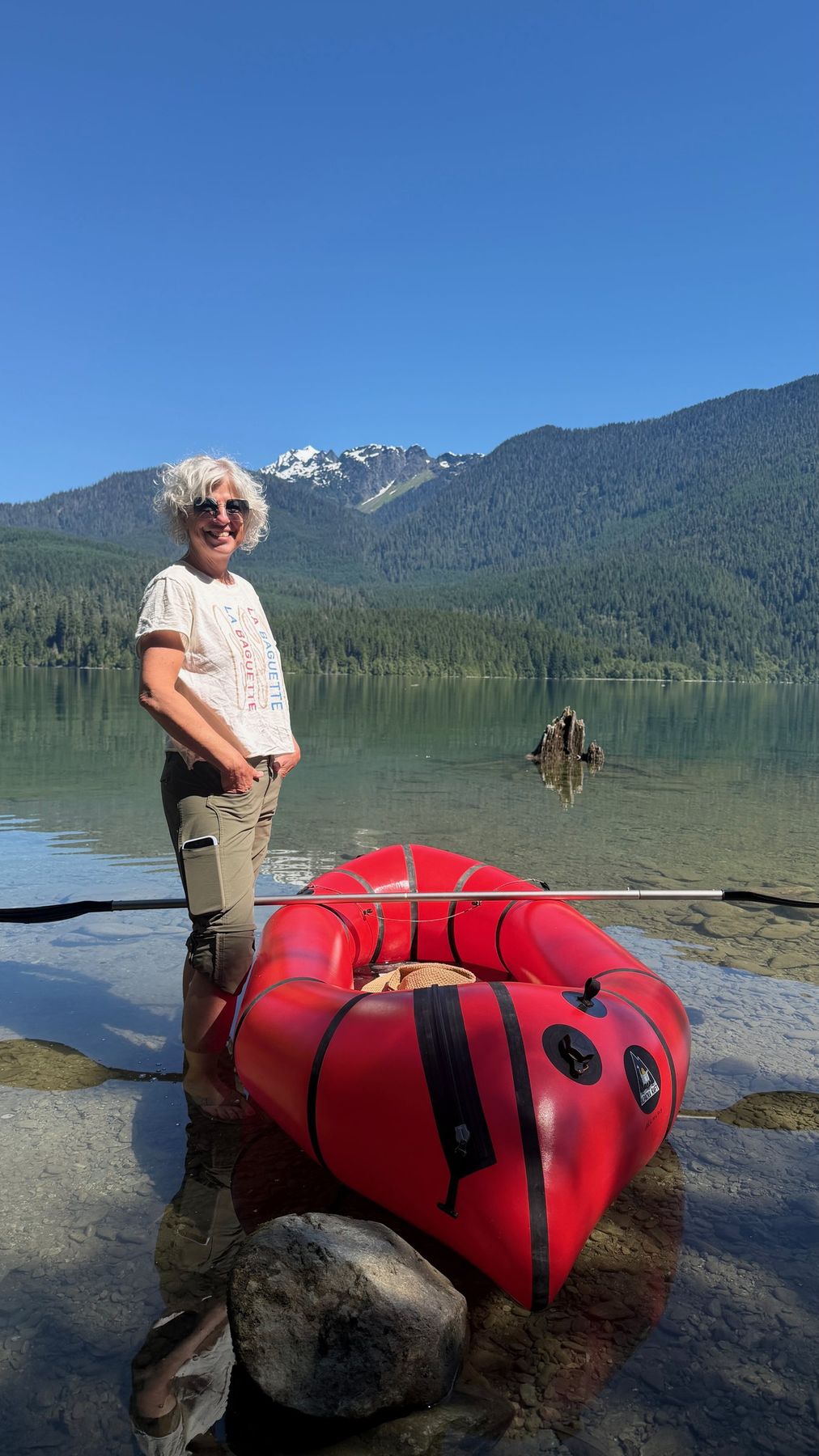 Suzanne smiling beside her red packraft on a clear mountain lake